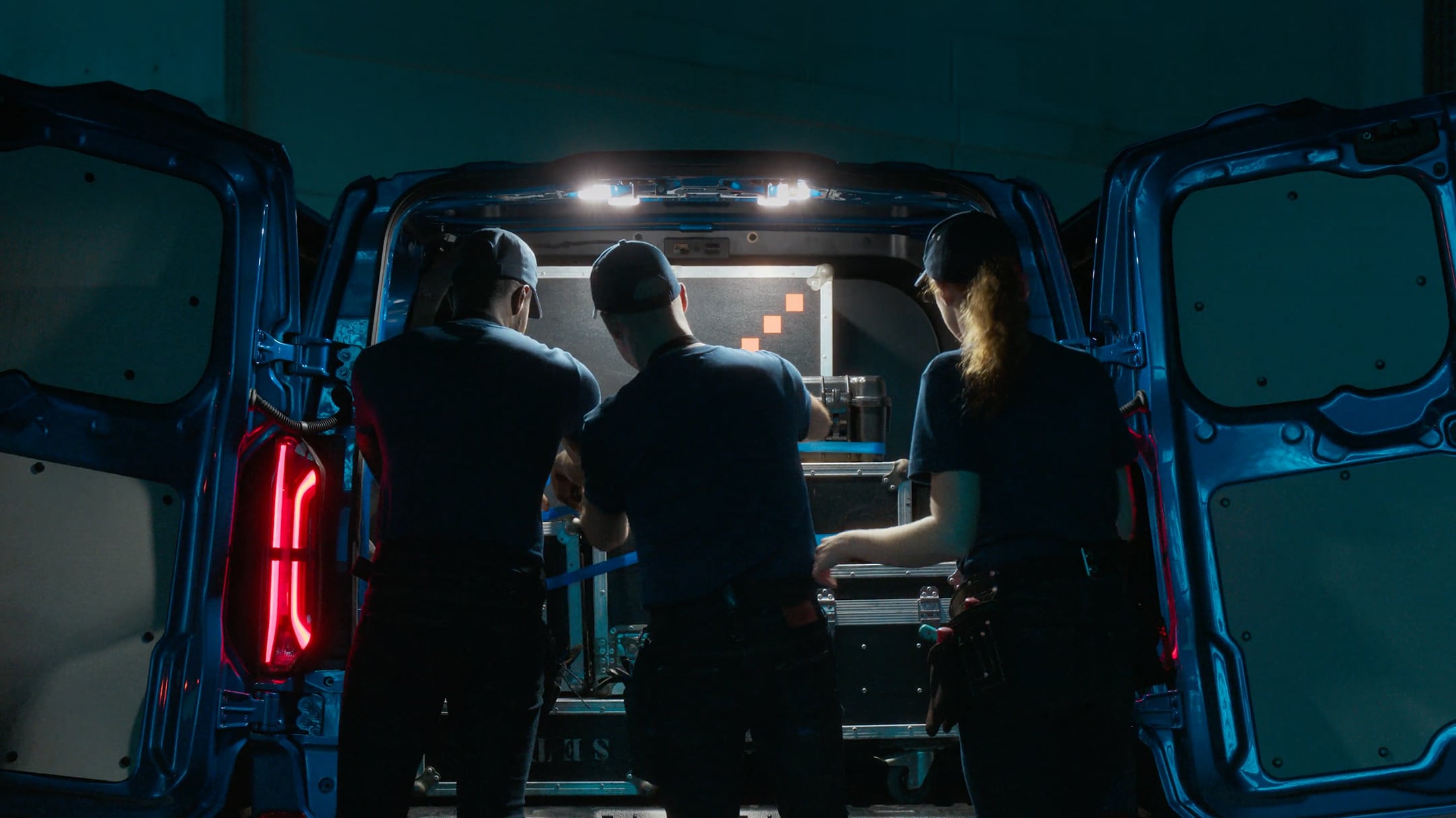 Ford Transit Custom Heckansicht. Drei Männer arbeiten bei geöffneter Hecktüre mit LED-Beleuchtung.