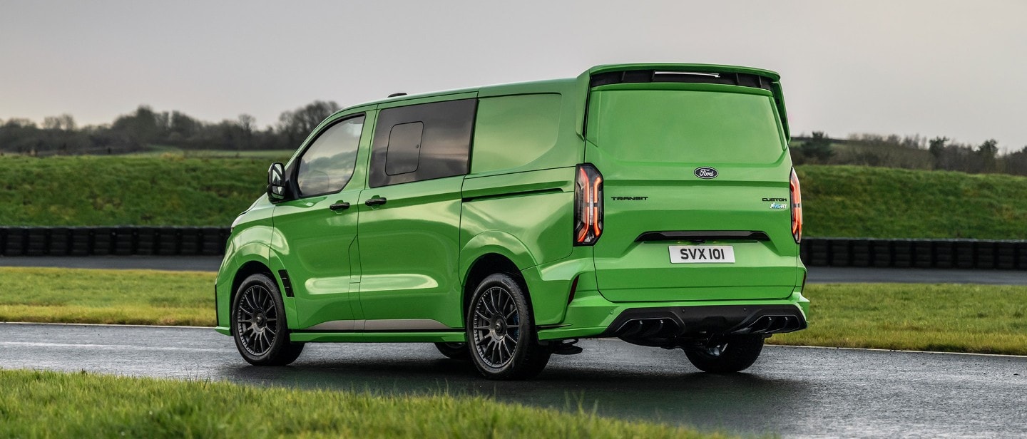 Ford Transit Custom MS-RT in Grün. Heckansicht, fahrend auf einer Straße. Landschaft im Hintergrund.