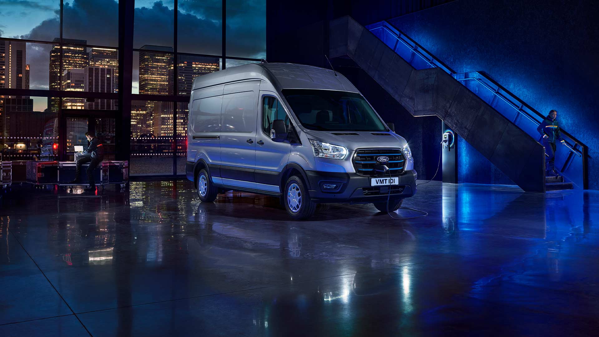 Ford E-Transit in Silber. Dreiviertelansicht, stehend in einer Halle. Skyline im Hintergrund.