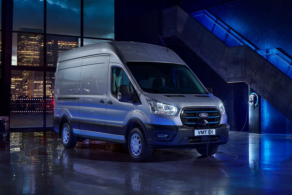 Ford E-Transit in Silber. Dreiviertelansicht in einer Halle mit großen Fenstern und Blick auf die nächtliche Skyline.