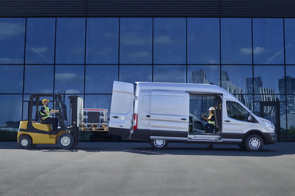 Ford E-Transit Kastenwagen in Silber. Seitenansicht, parkend vor modernem Gebäude mit geöffneter Schiebetür und Hecktür.