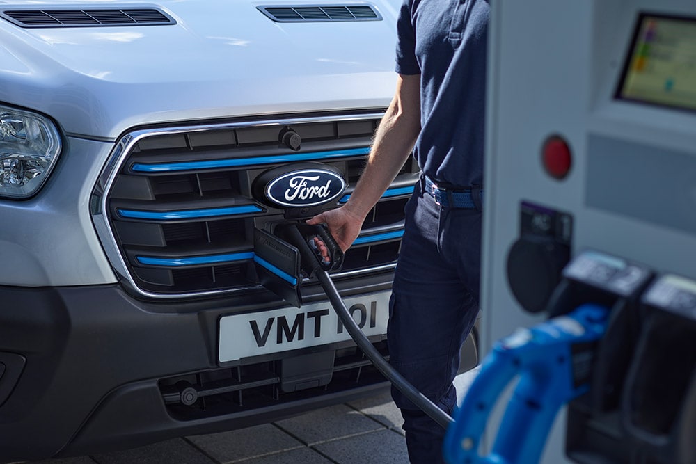 Ford E-Transit in Silber. Person schließt das Ladekabel an das Fahrzeug an.