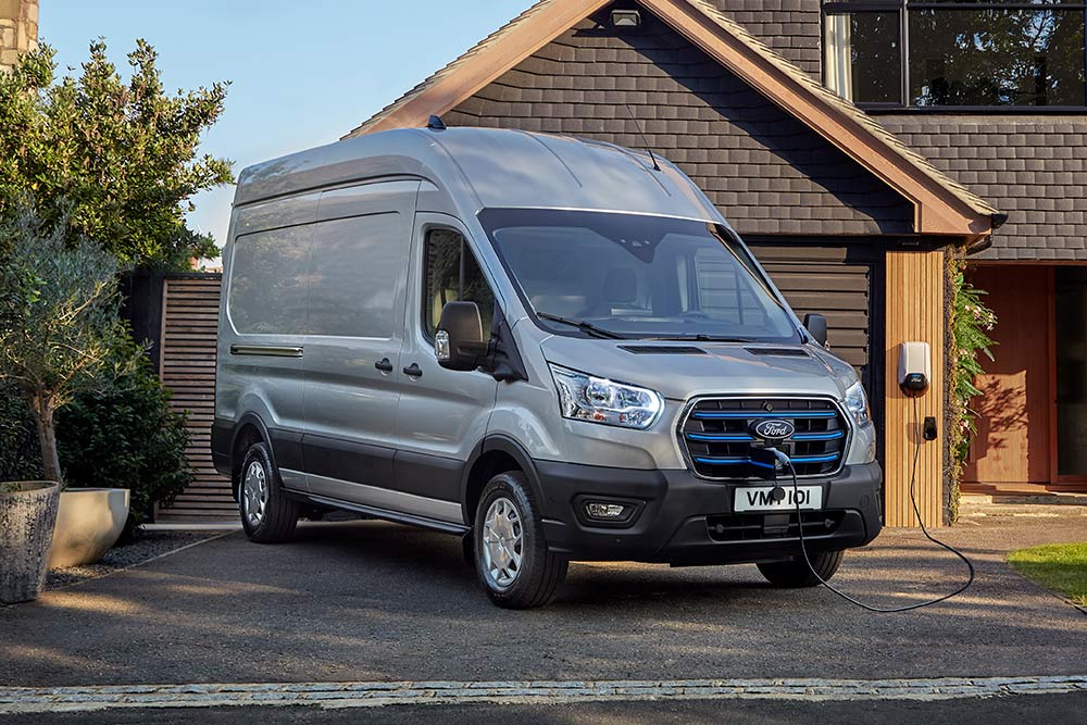 Ford E-Transit in Silber. Dreiviertelansicht, lädt an einer Wallbox vor der Garage eines Einfamilienhauses.