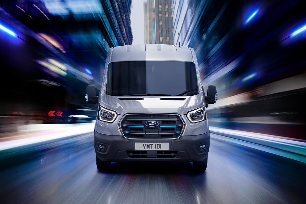Ford E-Transit in Silber. Frontansicht, fährt durch eine Innenstadt.