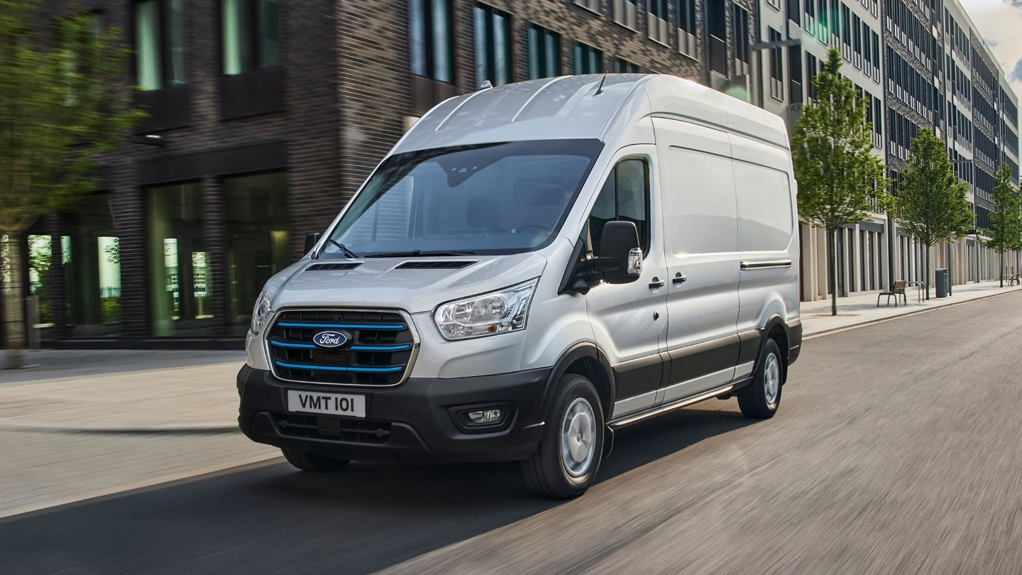 Ford E-Transit Kastenwagen in Silber. Dreiviertelansicht von vorne, fahrend entlang Hochhäusern