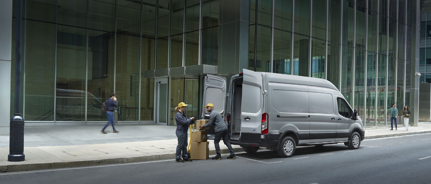 Ford E-Transit in Silber. Ansicht der geöffneten Hecktür, Frau und Mann beladen das Fahrzeug mit Kisten