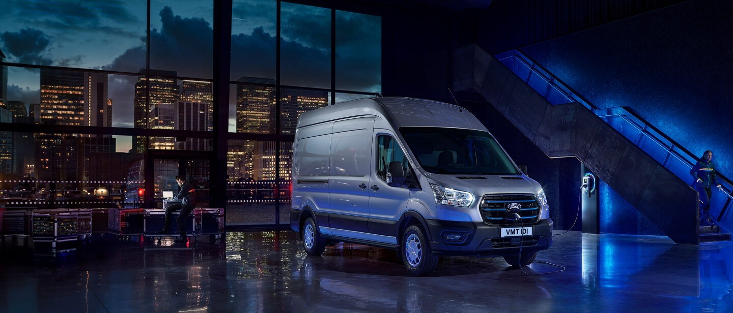 Ford E-Transit in Silber. Dreiviertelansicht, stehend in einer Halle. Skyline im Hintergrund.