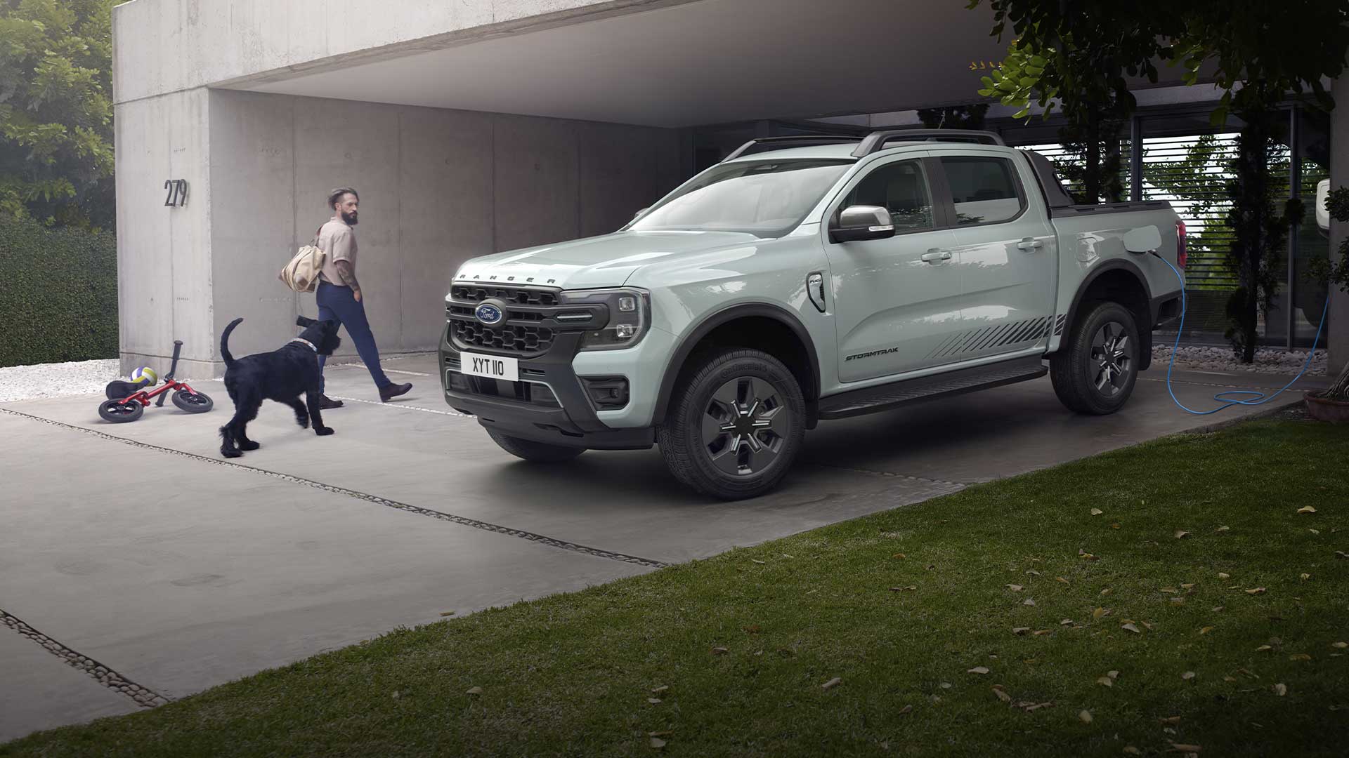 Ford Ranger Plug-in-Hybrid in Grau. Dreiviertelansicht, parkend vor einem Wohnhaus. Mann mit Hund gehen an Fahrzeug vorbei. 
