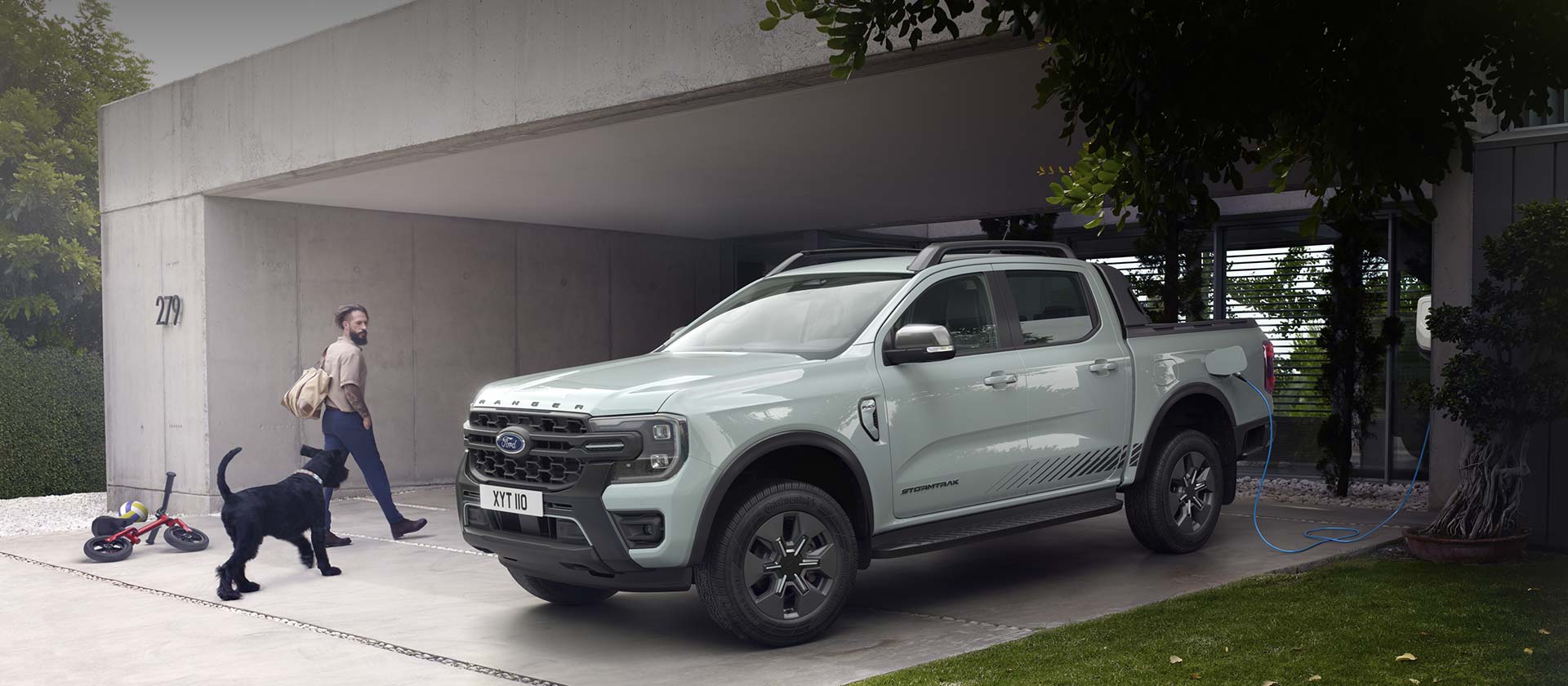 Ford Ranger Plug-in-Hybrid in Grau. Dreiviertelansicht, parkend vor einem Wohnhaus. Mann mit Hund gehen an Fahrzeug vorbei.