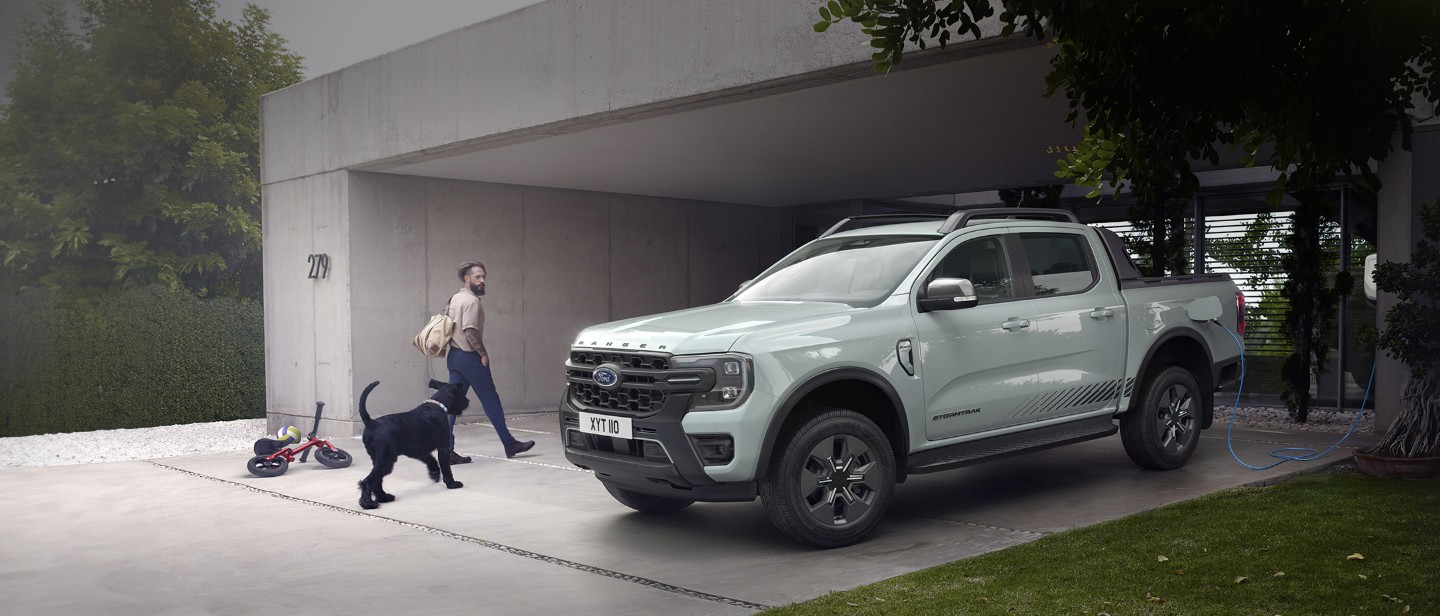 Ford Ranger Plug-in-Hybrid in Grau. Dreiviertelansicht, parkend vor einem Wohnhaus. Mann mit Hund gehen an Fahrzeug vorbei.