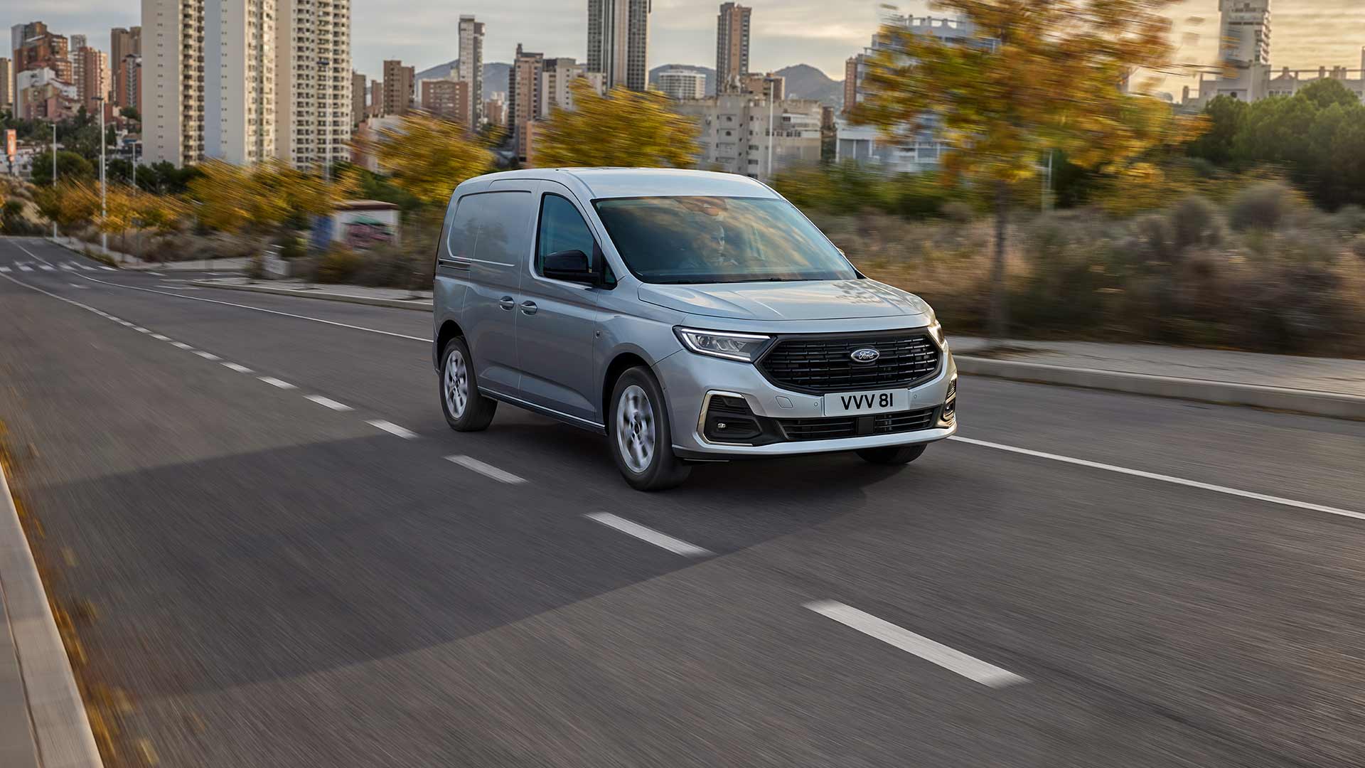 Ford Transit Connect in Silber. Dreiviertelansicht, fahrend auf einer Straße bei Sonnenuntergang. Stadt im Hintergrund. 