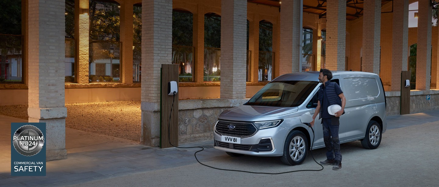 Ford Transit Connect in Silber. Dreiviertelansicht, parkend vor einer Halle. Mann schließt Fahrzeug an Ladestation an.
