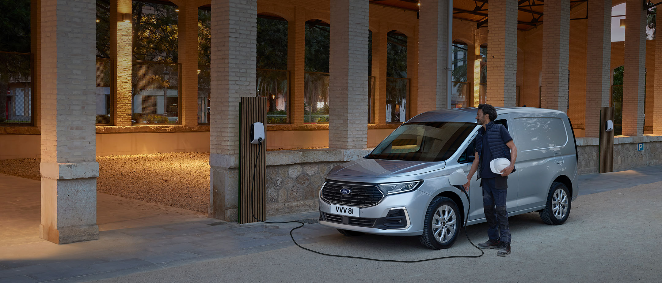 Ford Transit Connect in Silber. Dreiviertelansicht, parkend vor einer Halle. Mann schließt Fahrzeug an Ladestation an.