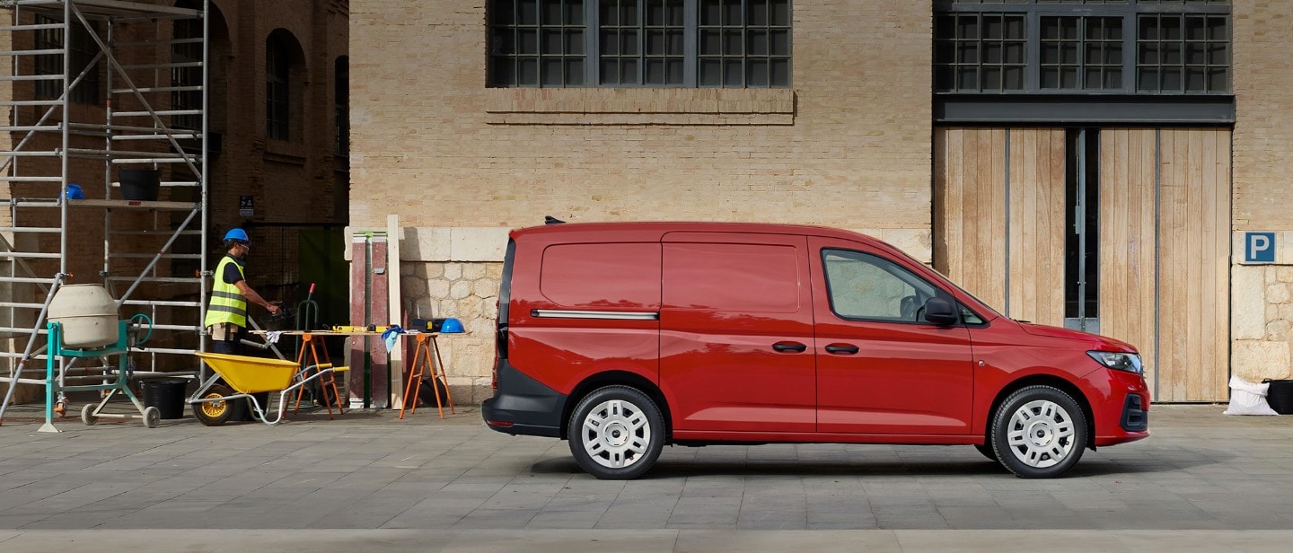 Ford Transit Connect in Rot. Seitenansicht, parkt vor einem Backsteingebäude mit Baustelle im Hintergrund.