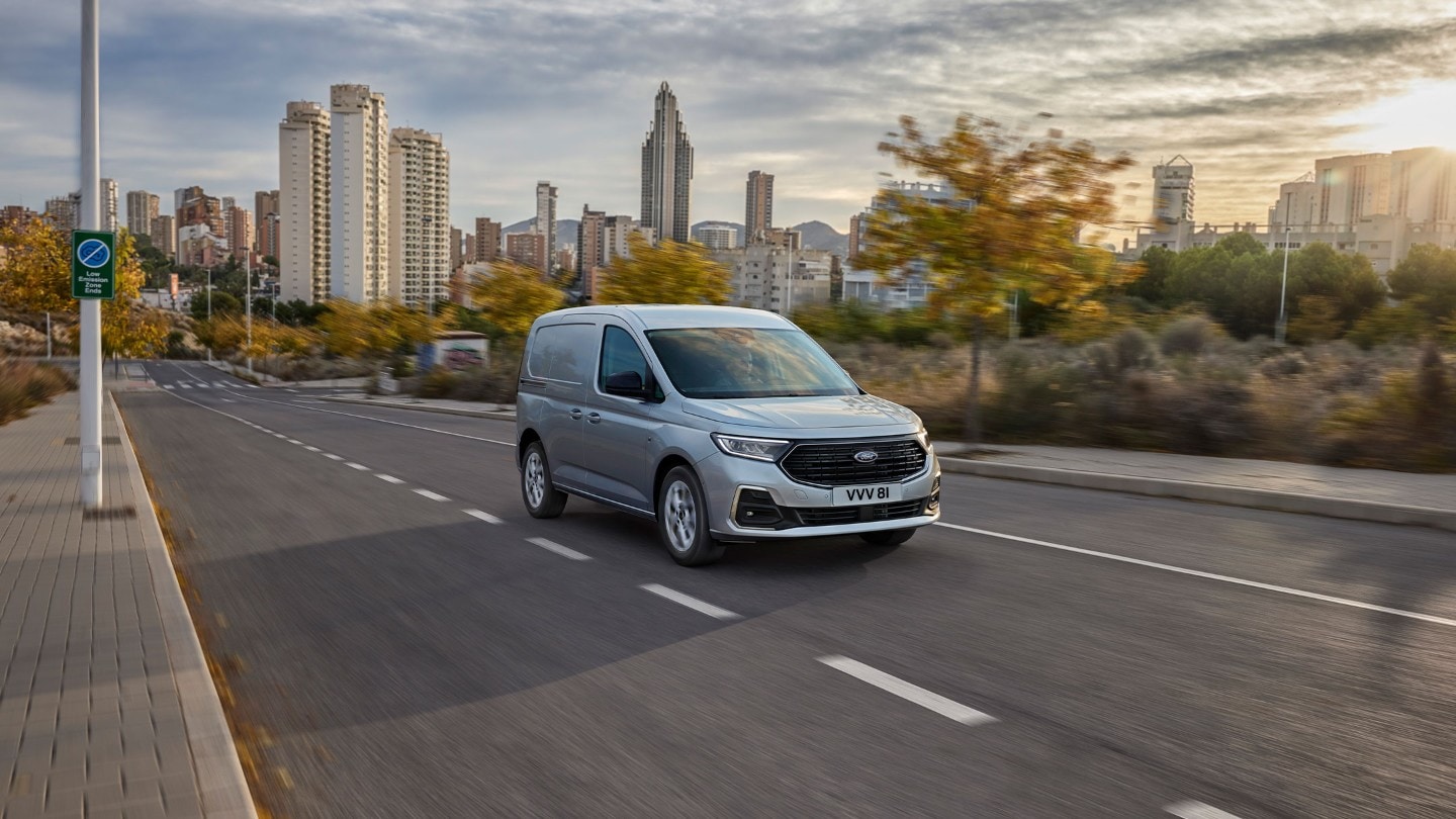 Ford Transit Connect in Silber. Frontansicht, fährt auf einer Straße mit Skyline im Hintergrund.