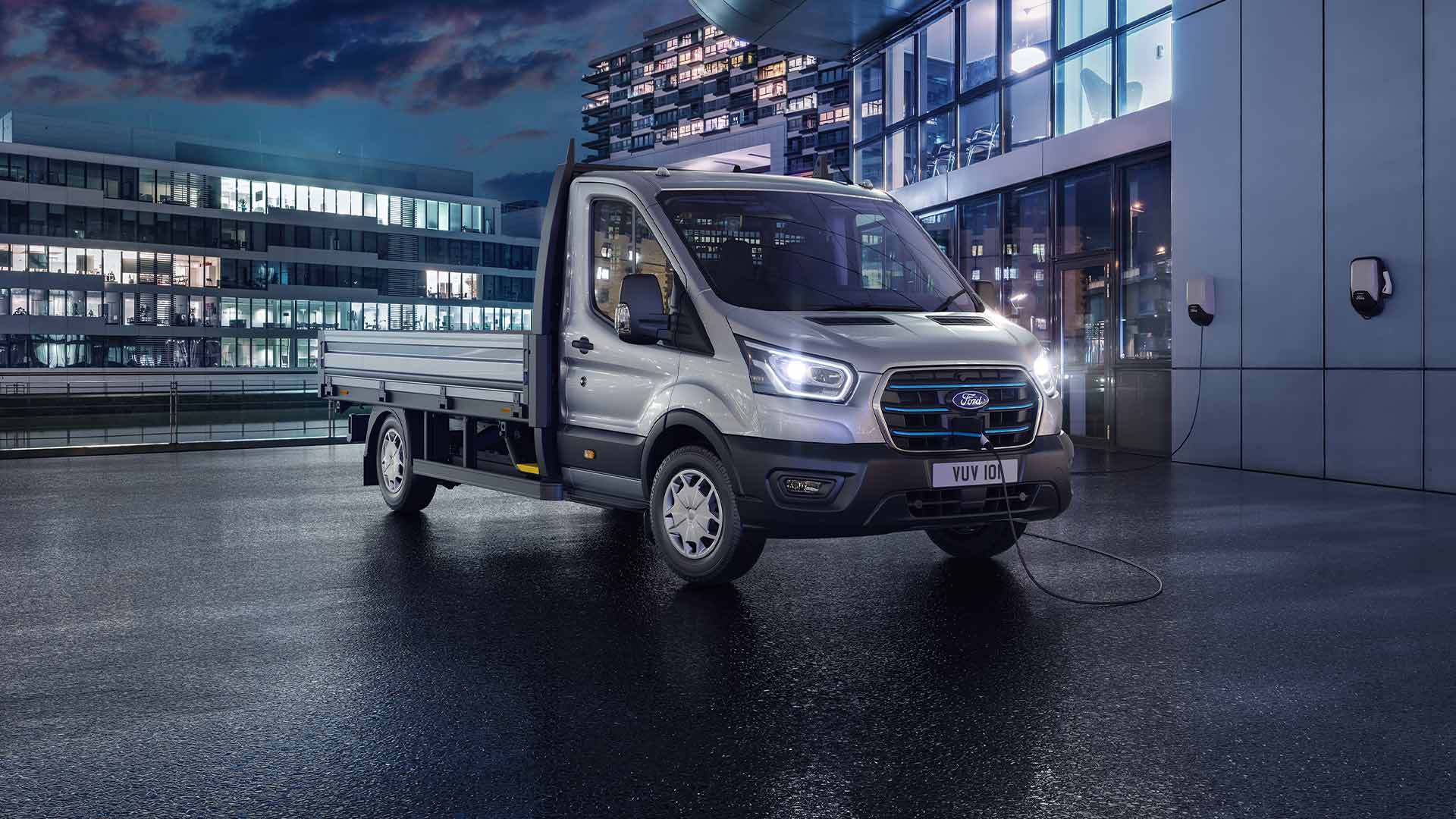 Ford E-Transit Pritschenwagen in Silber. Dreiviertelansicht bei Nacht vor beleuchteten Gebäuden.