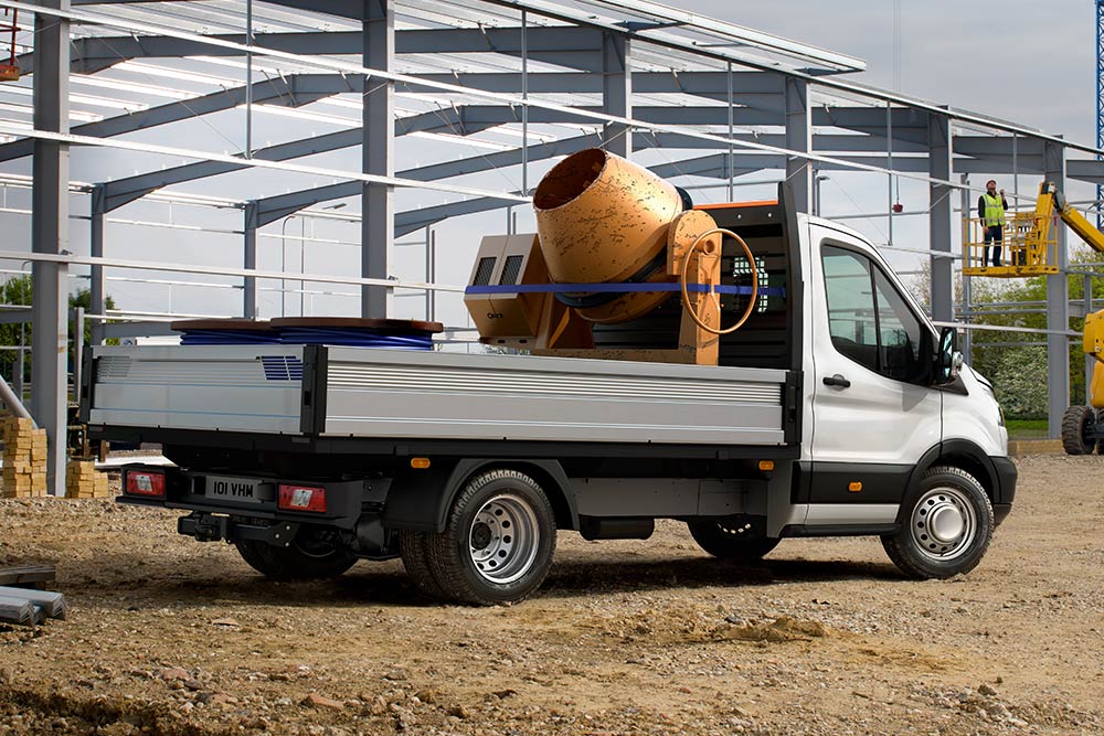 Ford Transit Pritschenwagen mit Einzelkabine in Weiß. Dreiviertelansicht, auf einer Baustelle, mit Betonmischer auf der Ladefläche. 