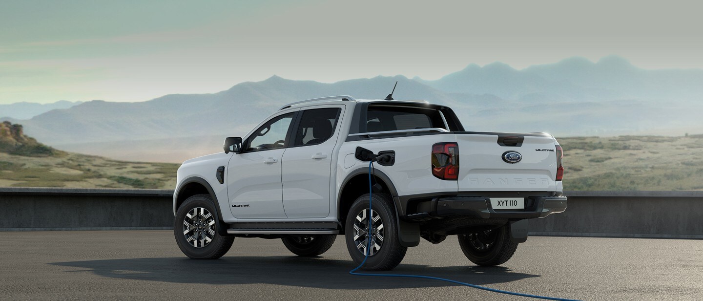 Ford Ranger Plug-in-Hybrid in Weiß. Dreiviertelansicht, an einer Ladestation angeschlossen mit Bergen im Hintergrund.