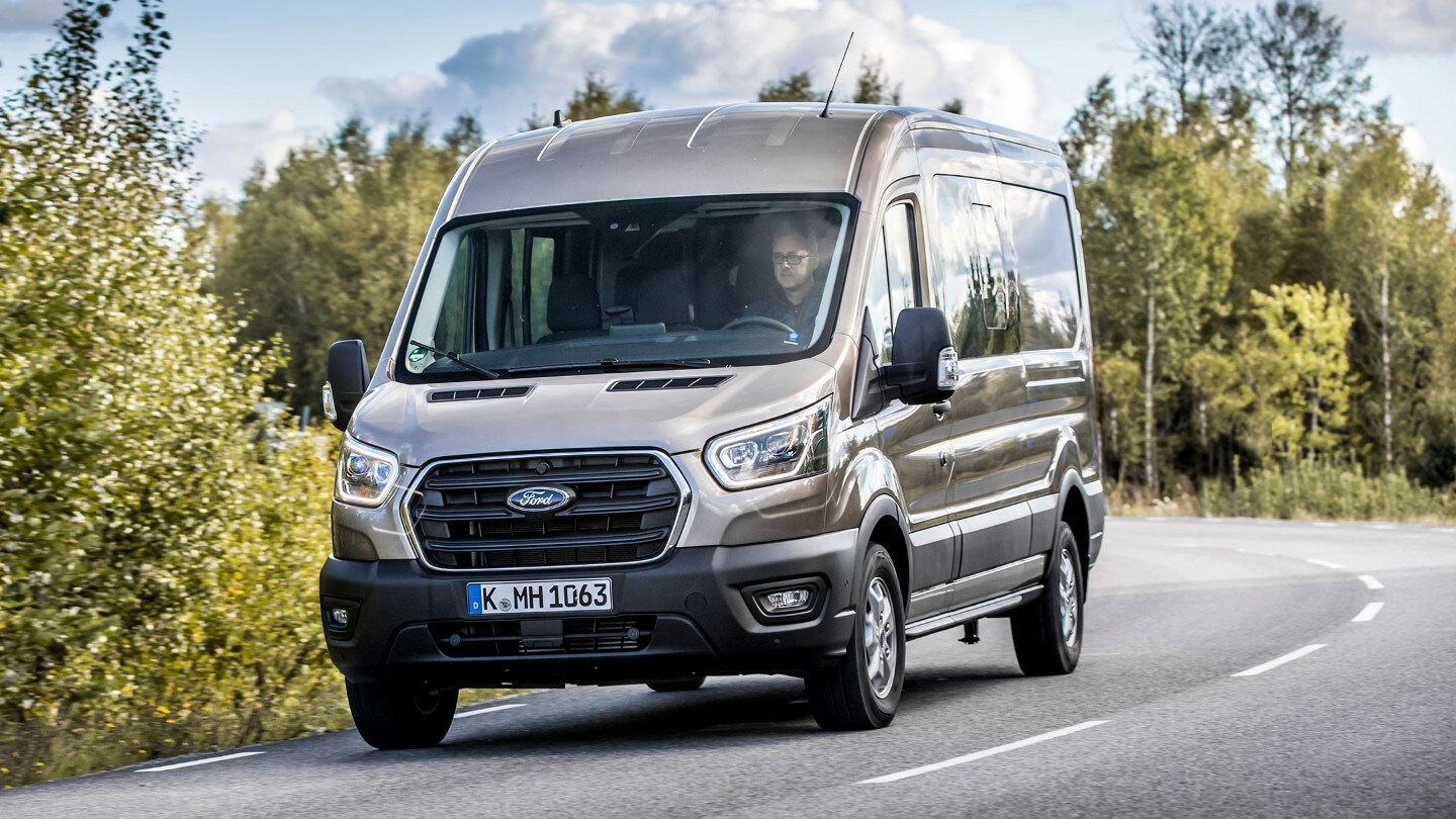 Ford Transit Doppelkabine in Silber. Dreiviertelansicht von vorne, fahrend auf einer Landstraße