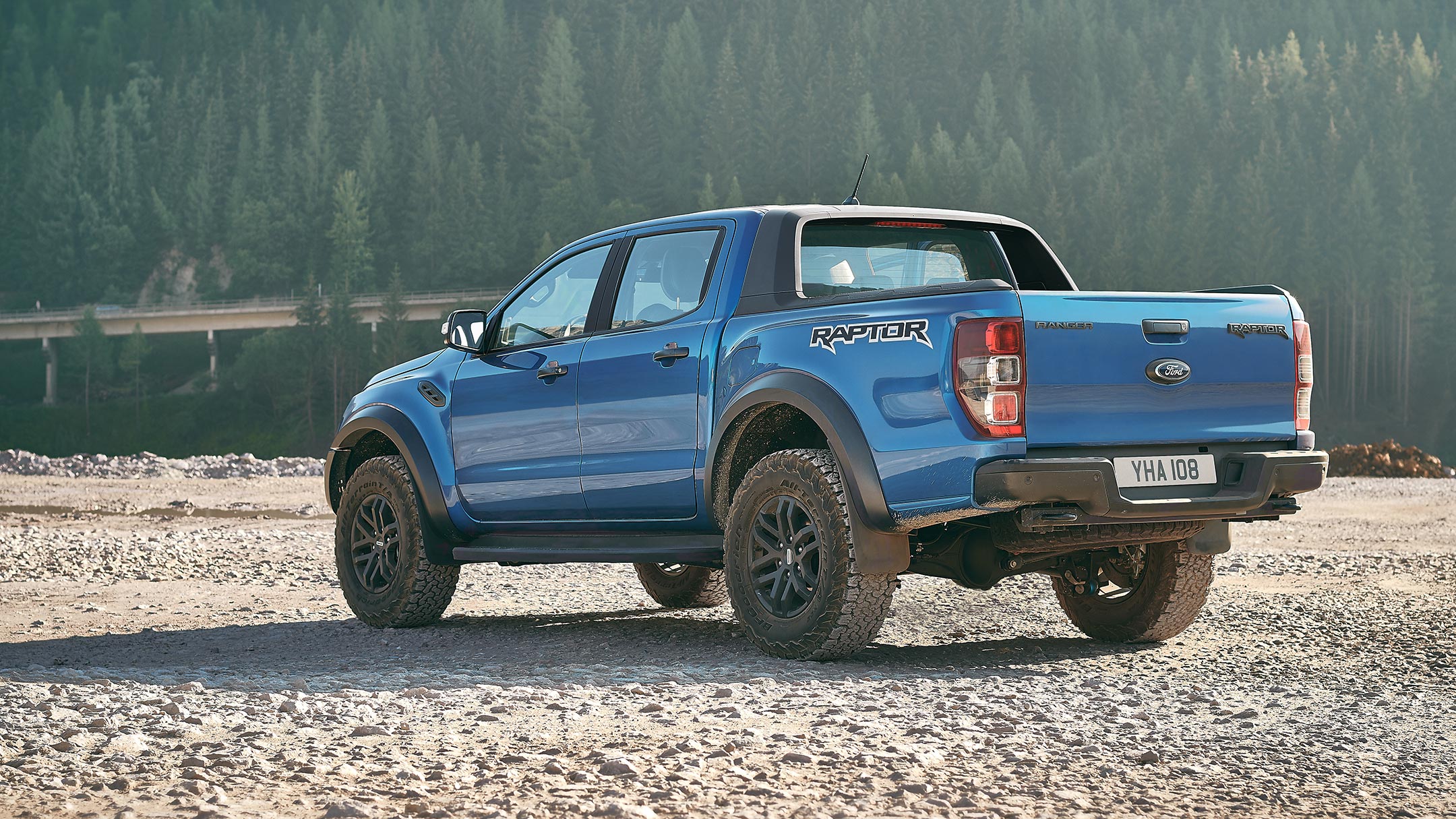 Ford Ranger Raptor in Blau. Dreiviertelansicht von hinten, parkend vor einem Wald