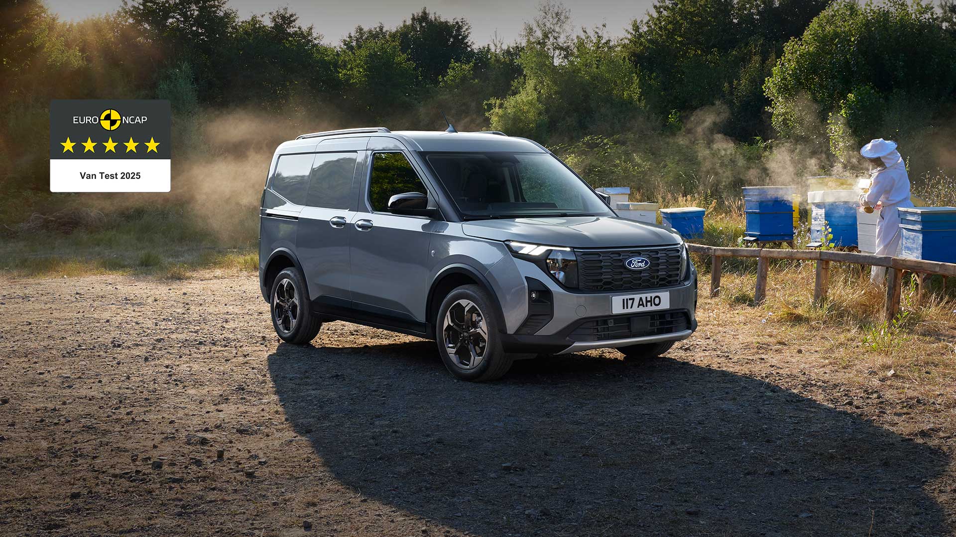 Ford Transit Courier in Silber. Dreiviertelansicht, parkend auf einem Gelände mit Bäumen im Hintergrund.