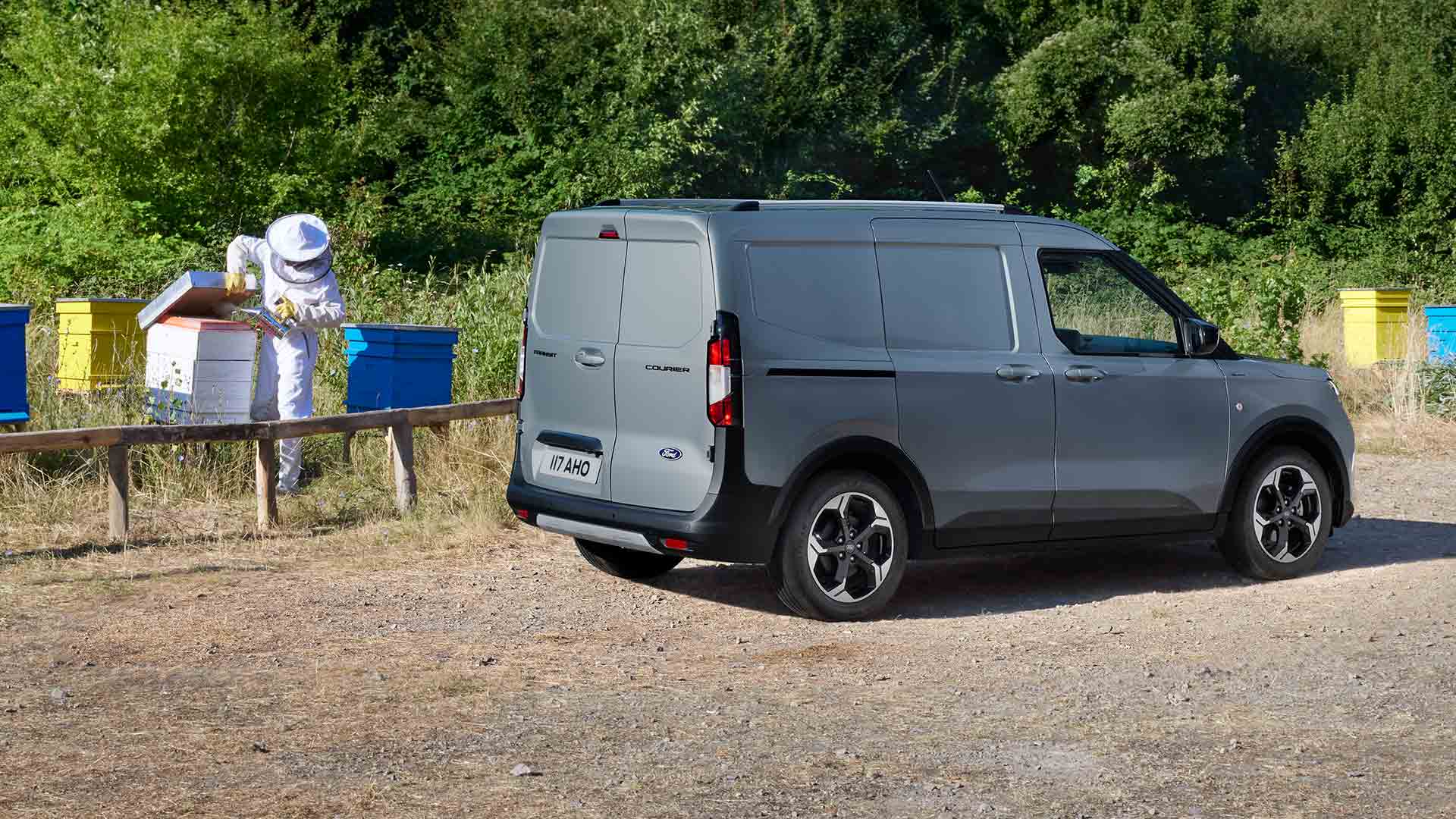Ford Transit Courier in Silber. Dreiviertelansicht, parkend auf einem Gelände mit Bäumen im Hintergrund. 