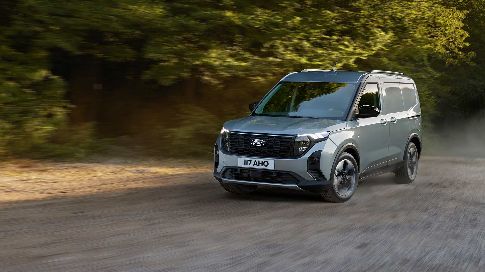 Ford Transit Courier in Silber. Dreiviertelansicht, fahrend auf einer unbefestigten Straße mit Bäumen im Hintergrund.