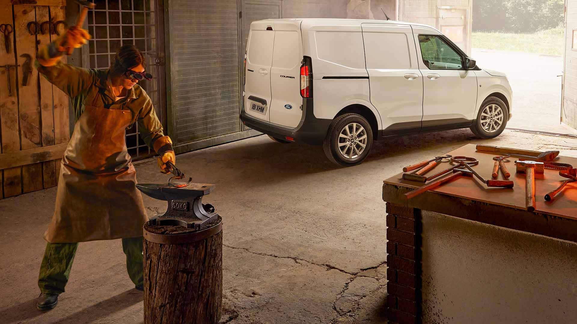 Ford Transit Courier in Weiß. Dreiviertelansicht, steht in einer Werkstatt. Schmied arbeitet im Vordergrund.