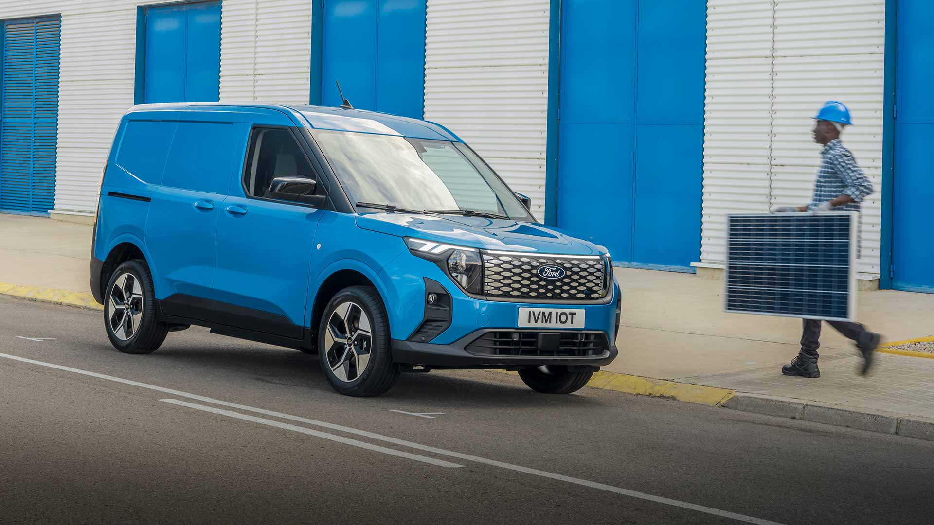 Ford E-Transit Courier in Blau. Dreiviertelansicht, parkend vor einem Gebäude. Mann nähert sich dem Fahrzeug.