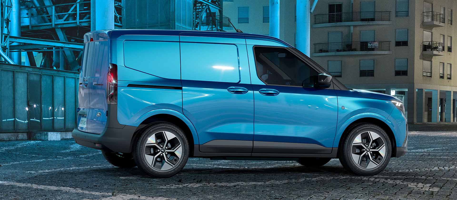 Ford E-Transit Courier in Blau. Seitenansicht, stehend vor einer Garage bei Nacht. 