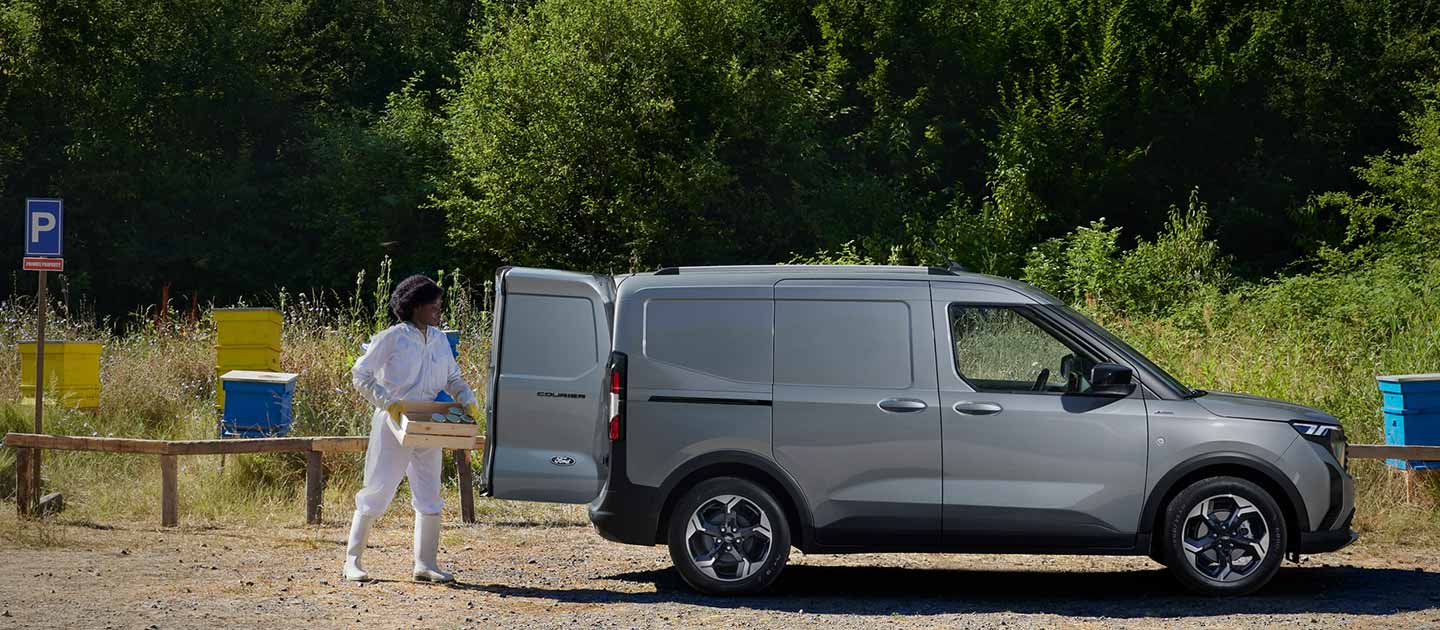 Ford Transit Courier in Silber. Seitenansicht, stehend auf einem Gelände mit Bäumen im Hintergrund und geöffneter Hecktür
