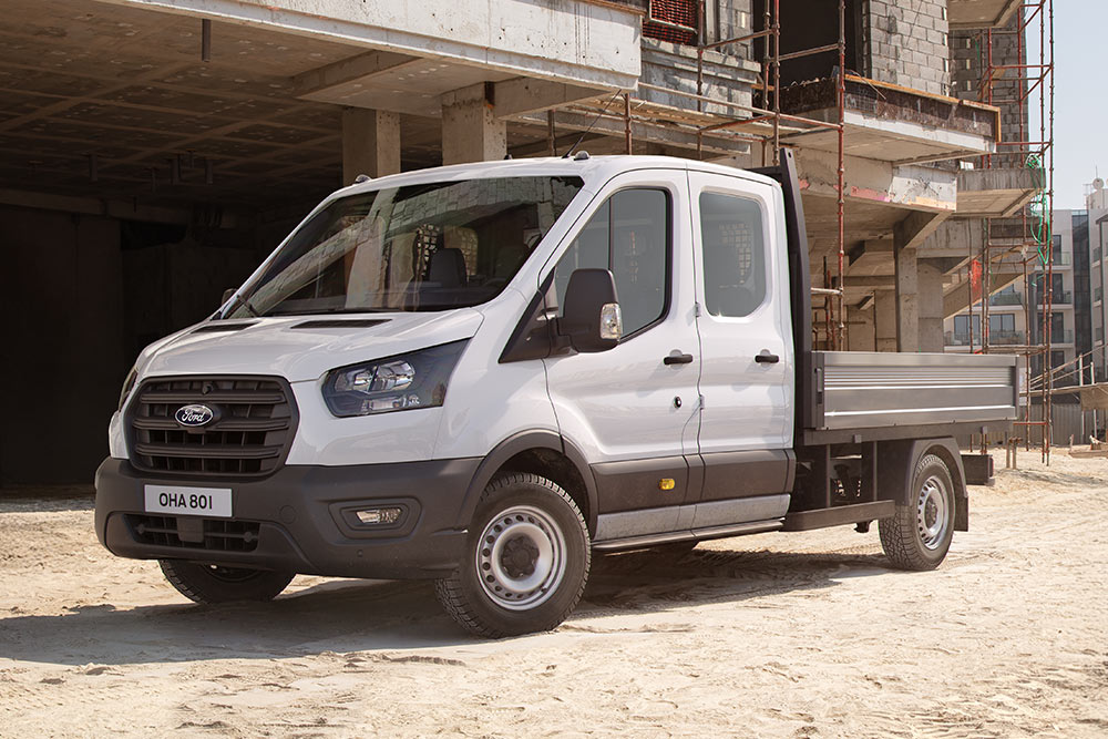 Ford Transit Pritschenwagen in Weiß. Dreiviertelansicht, parkt auf einer Baustelle.
