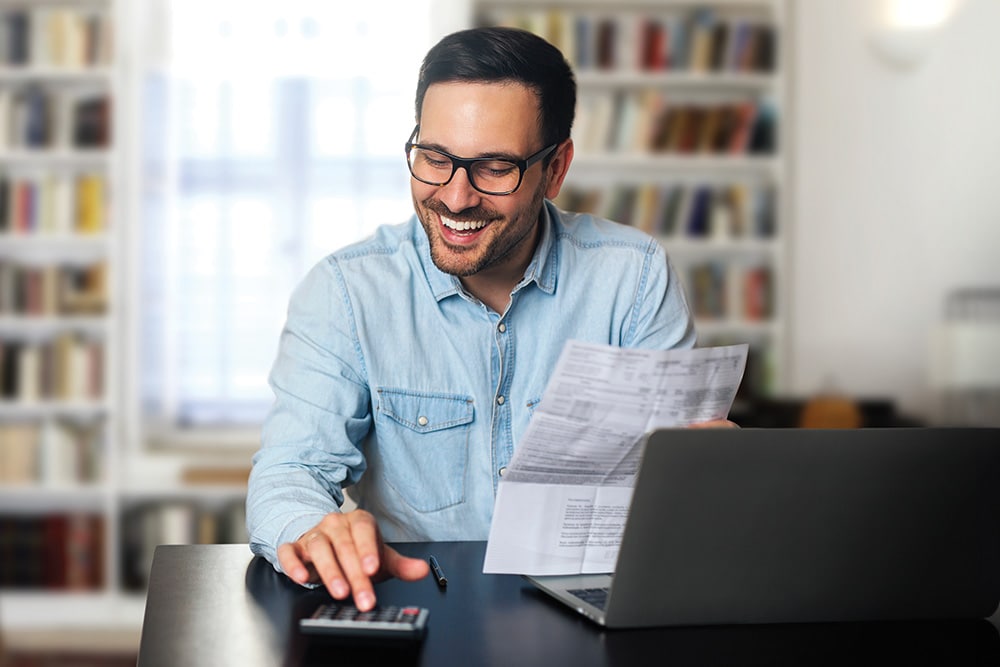 A man in glasses smiles while using a calculator and holding documents at a desk with a laptop.