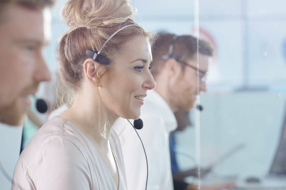 Drei Kundendienstmitarbeitende mit Headsets in einem Büro, eine lächelnde Frau im Vordergrund.