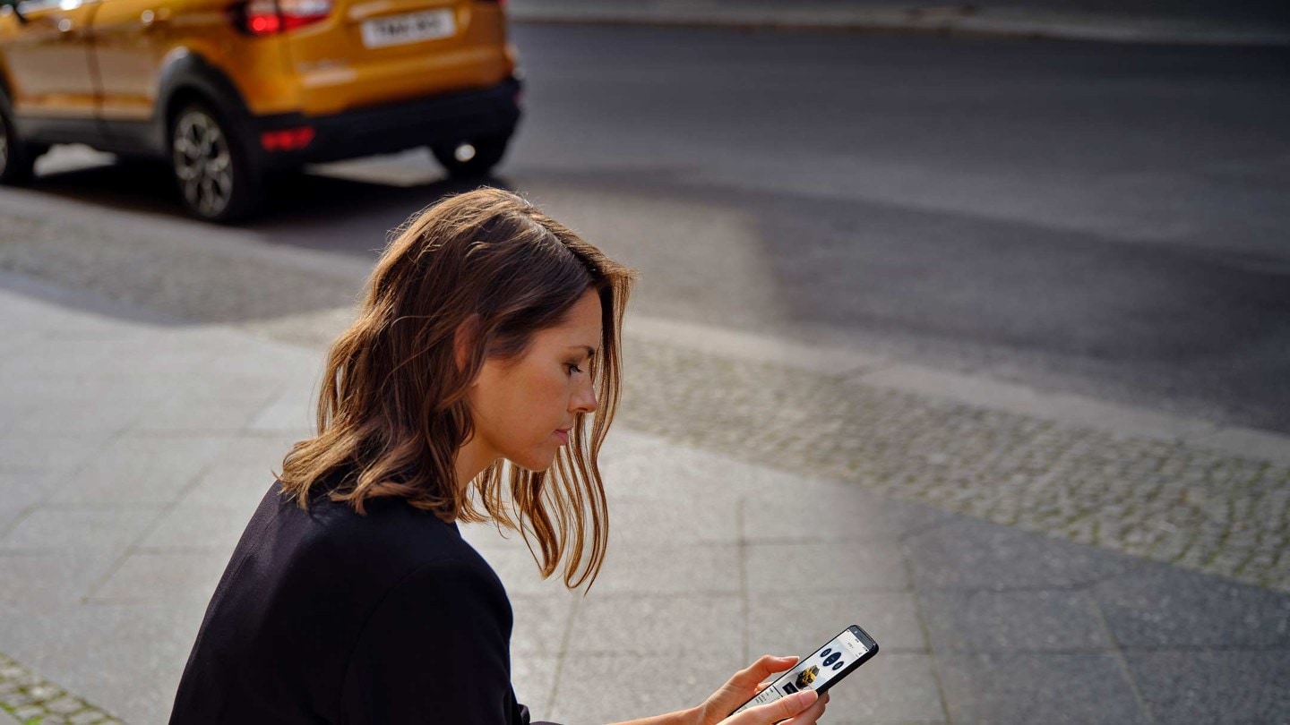 Frau mit einem Mobiltelefon in der Hand, neben ihr ein Ford EcoSport