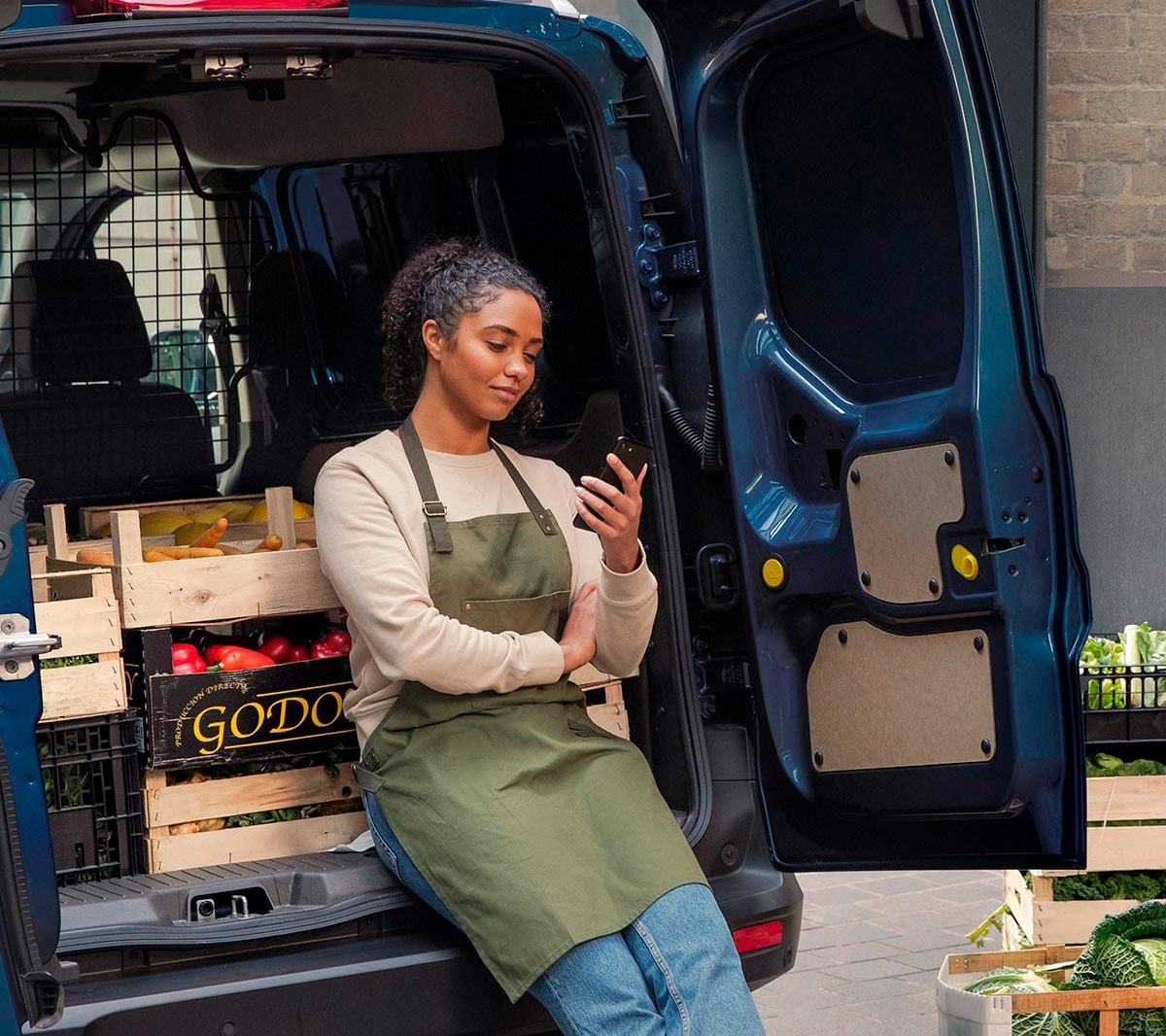 Frau schaut auf ihr Handy und lehnt an der Ladefläche eines Ford Transit Couriers