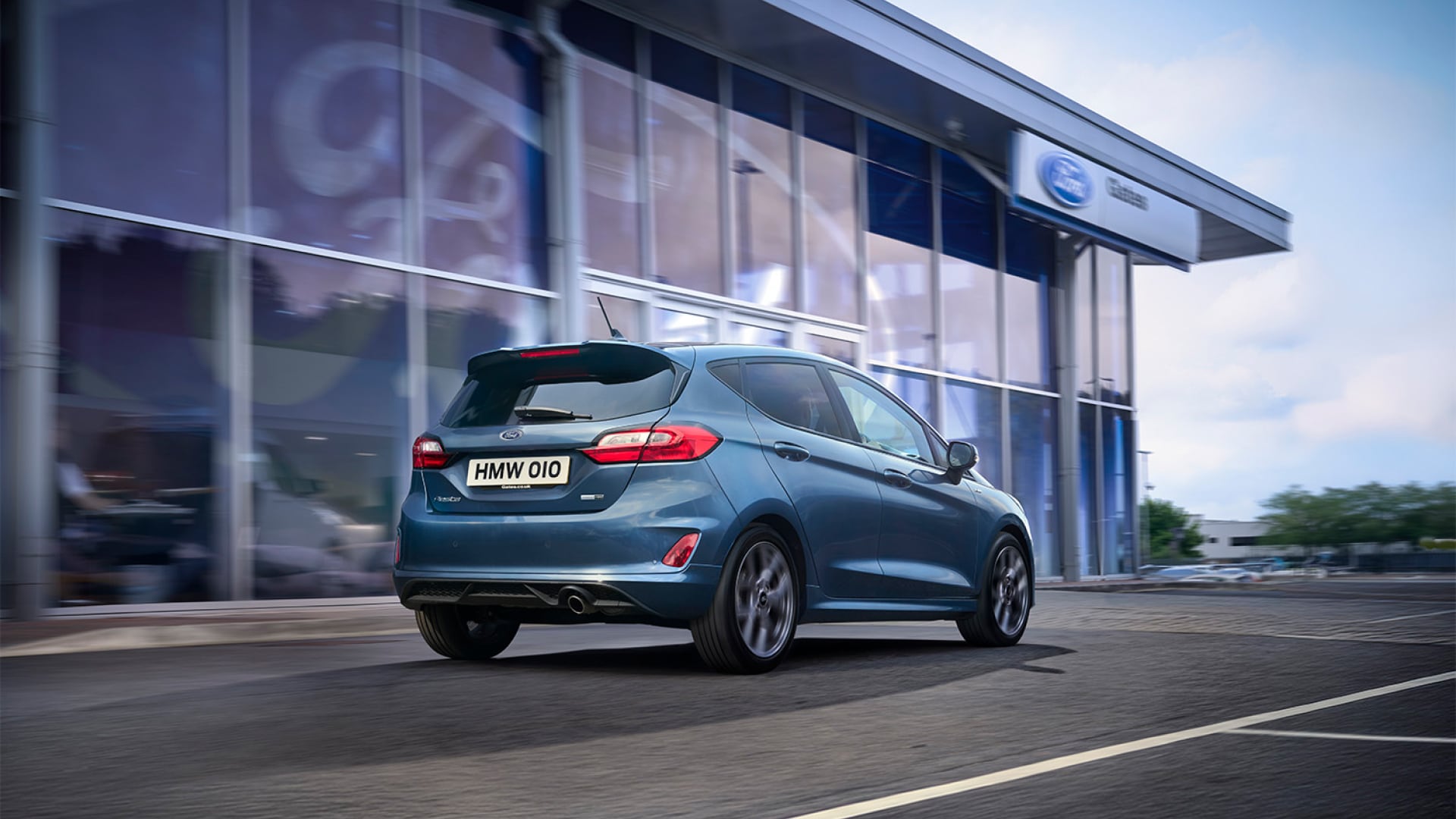 Ein blauer Kleinwagen fährt an einem Ford-Autohaus vorbei.