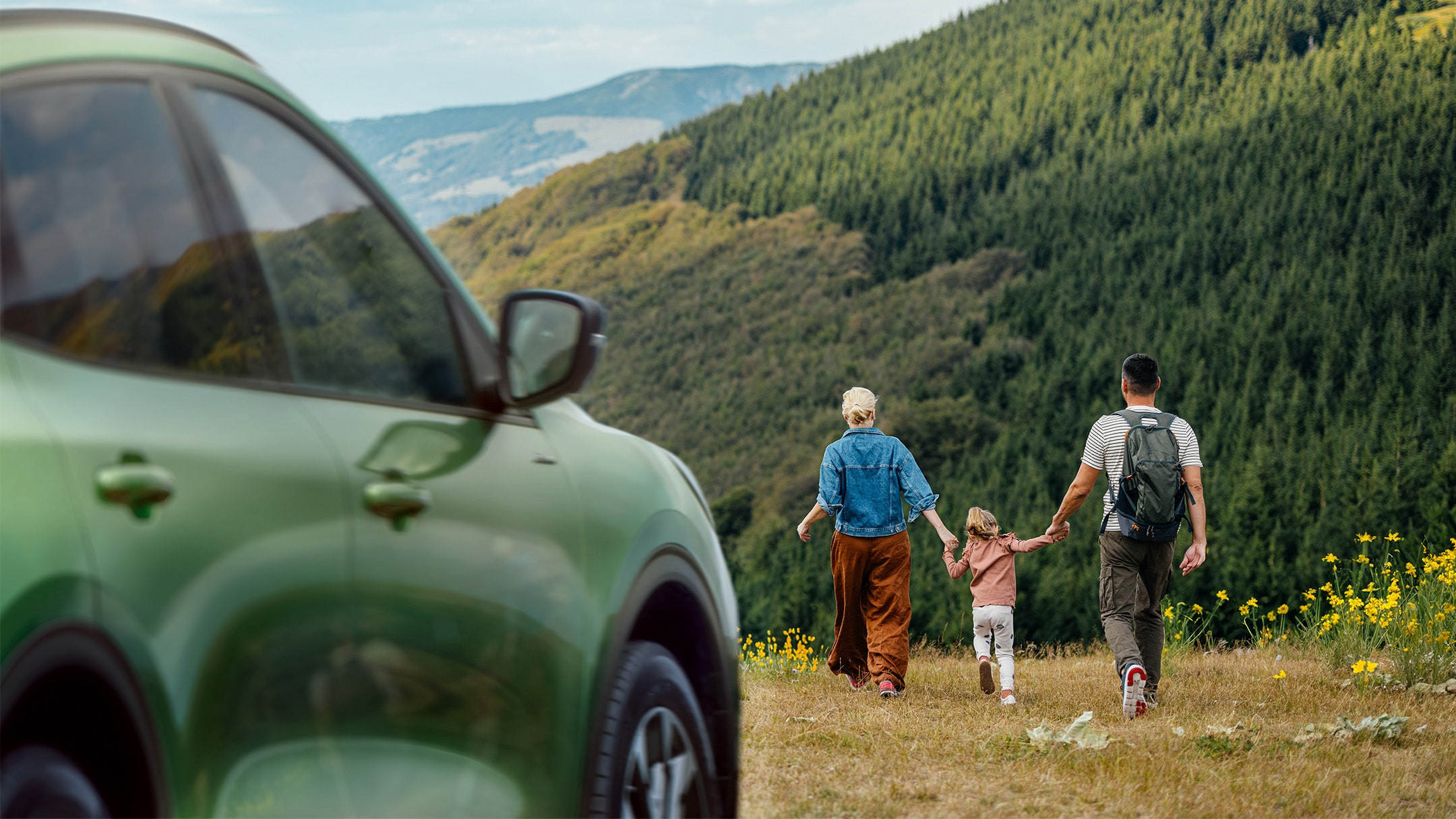 Ford Fahrzeug steht inmitten der Natur. Eine Familie spaziert fröhlich im Hintergrund über eine Wiese.