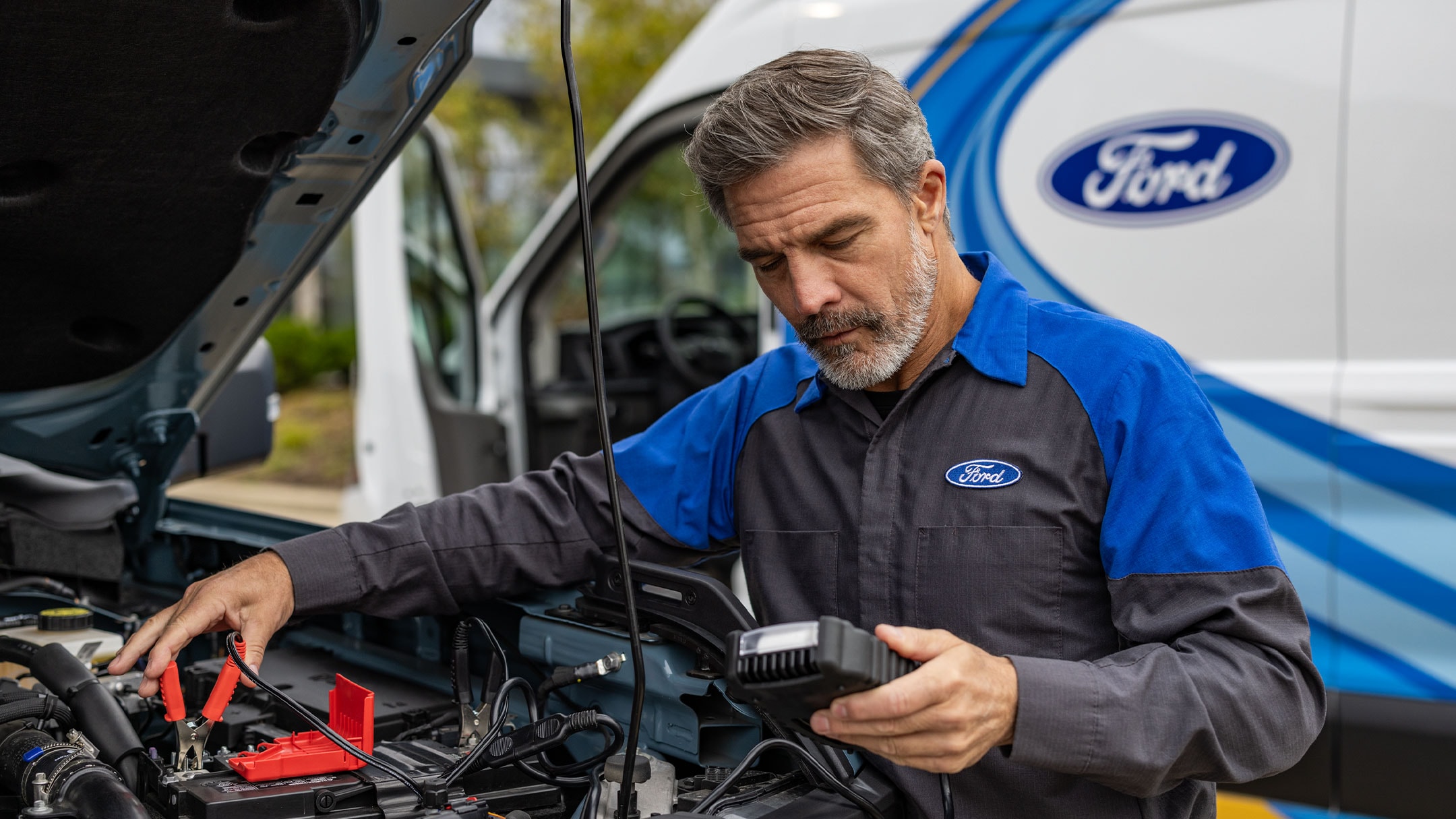  Ford-Techniker in Uniform schließt Überbrückungskabel an eine Autobatterie an, mit einem Ford-Service-Transporter in der Nähe.