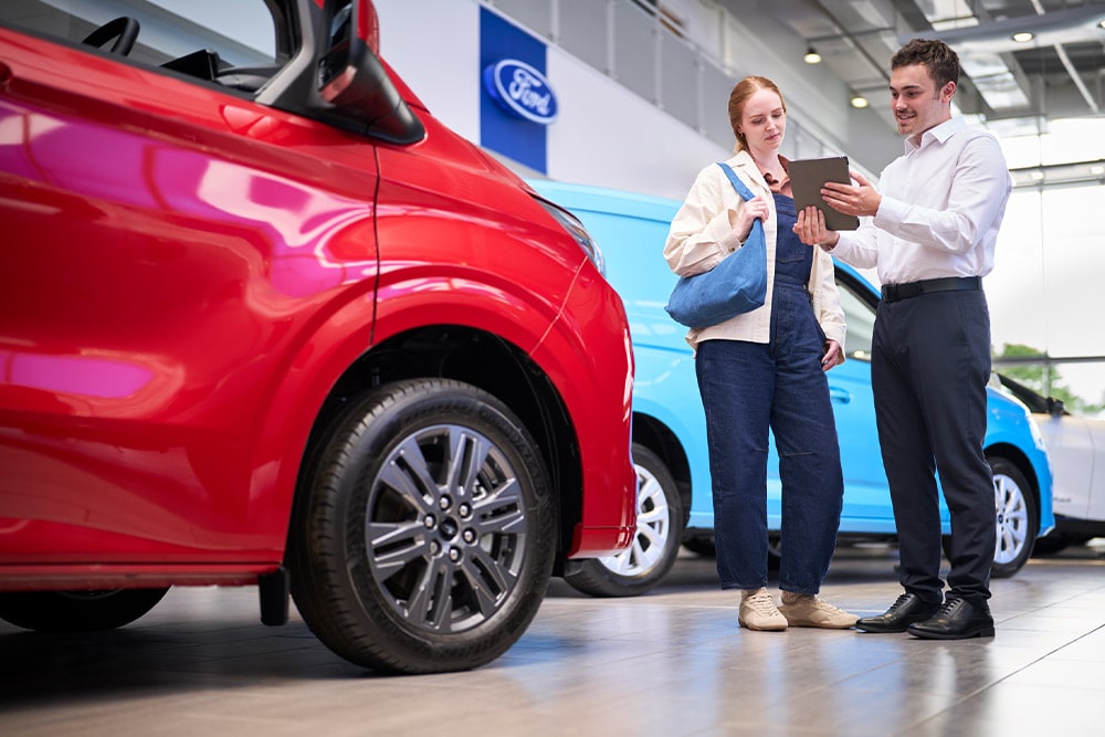 Ein Ford-Mitarbeiter zeigt einem Kunden in einem Ausstellungsraum mit roten und blauen Ford-Fahrzeugen ein Tablet. 