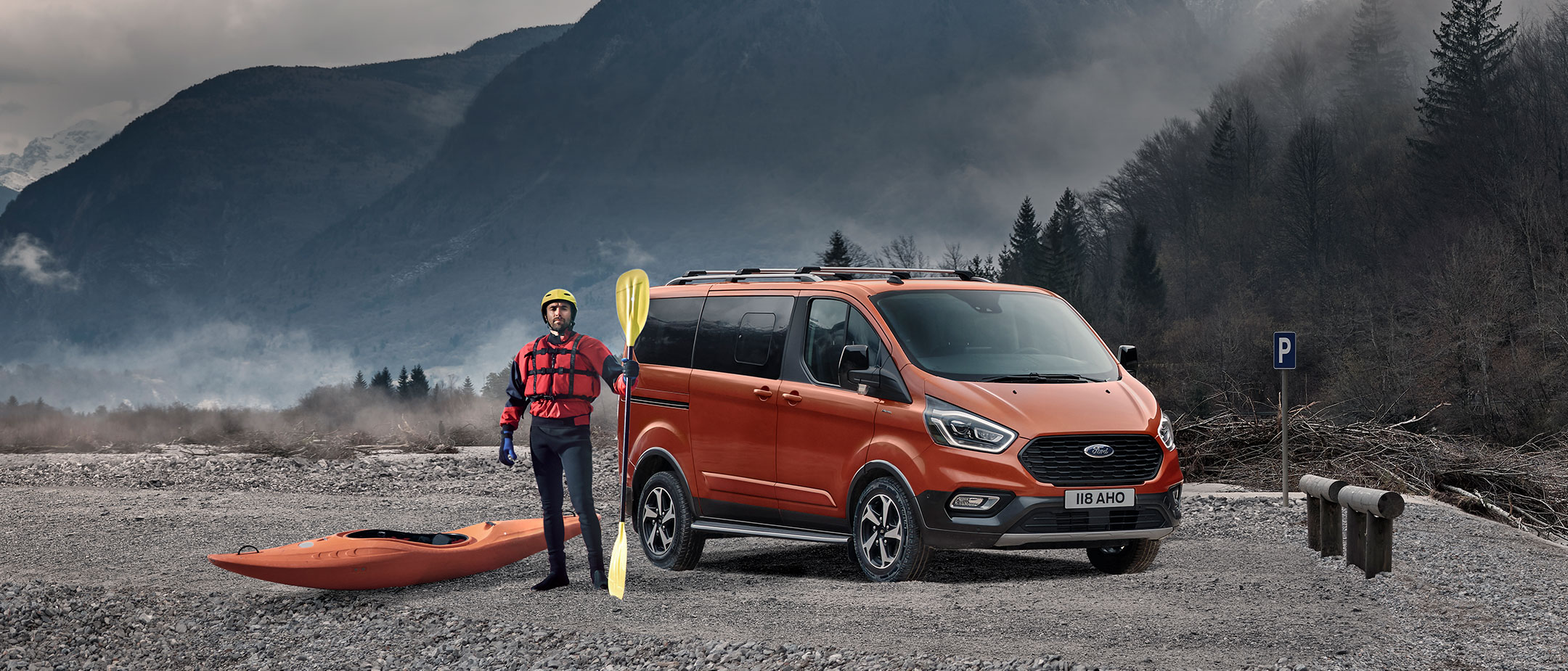 Ford Tourneo Custom in Orange. Dreiviertelansicht, steht auf Kies mit Bergen im Hintergrund