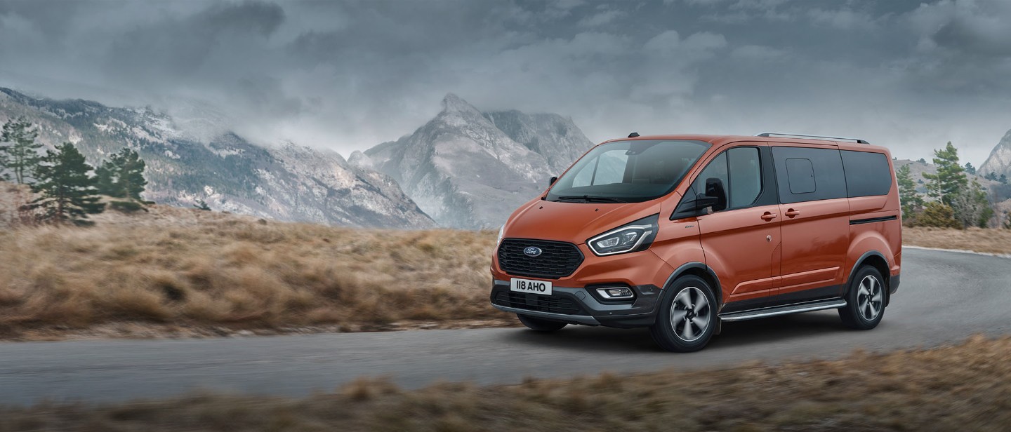 Ford Tourneo Custom in Orange. Dreiviertelansicht, auf einer Straße in den Bergen